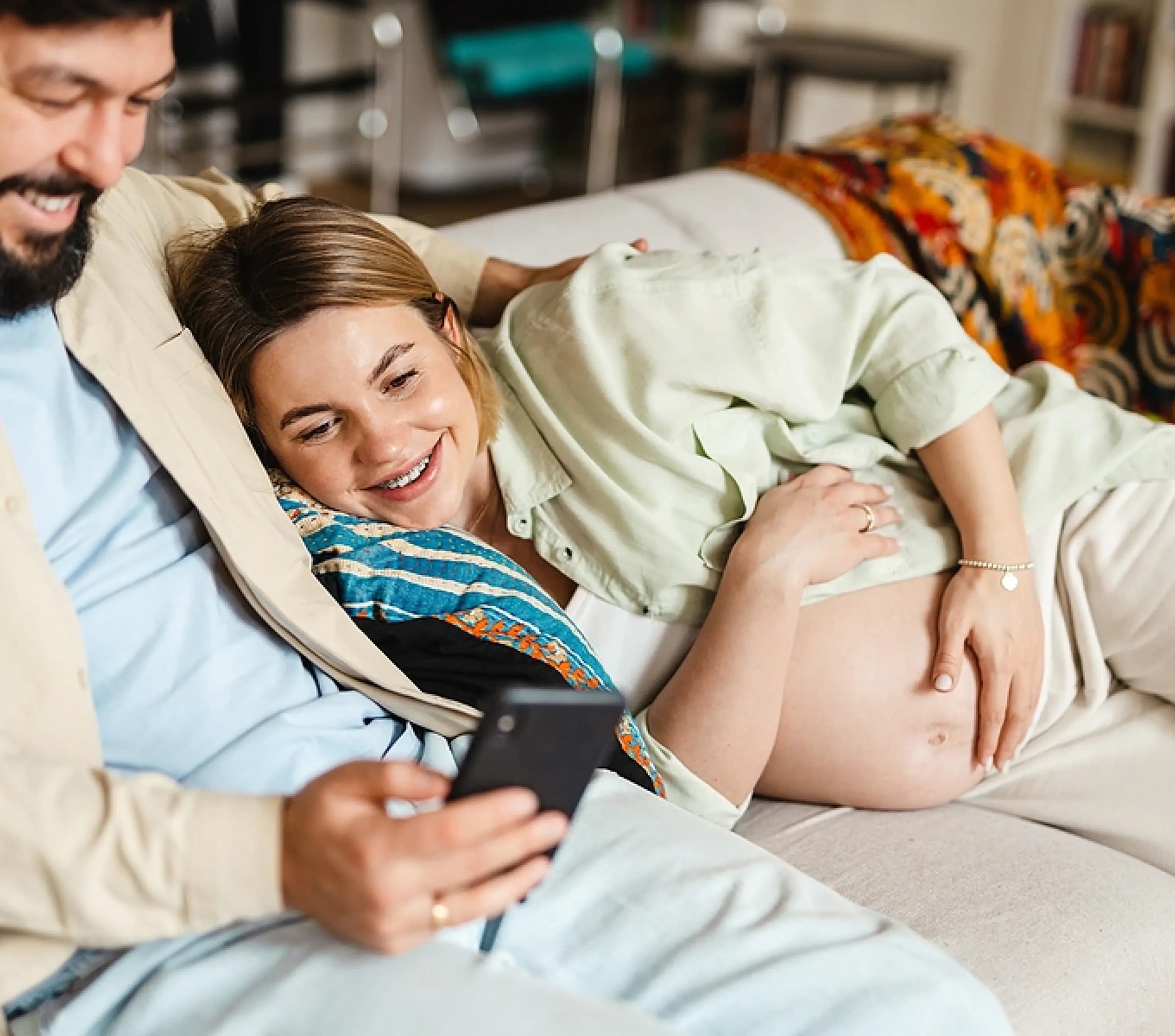 Lachende zwangere vrouw en haar partner volgen samen een zwangerschapscursus op hun telefoon, thuis op de bank